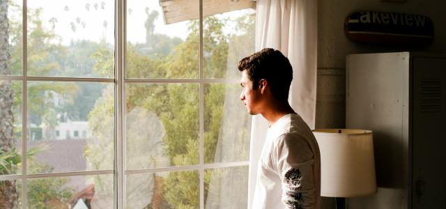 man wearing gray and black crew-neck shirt standing and looking out window by Hamish Duncan courtesy of Unsplash.